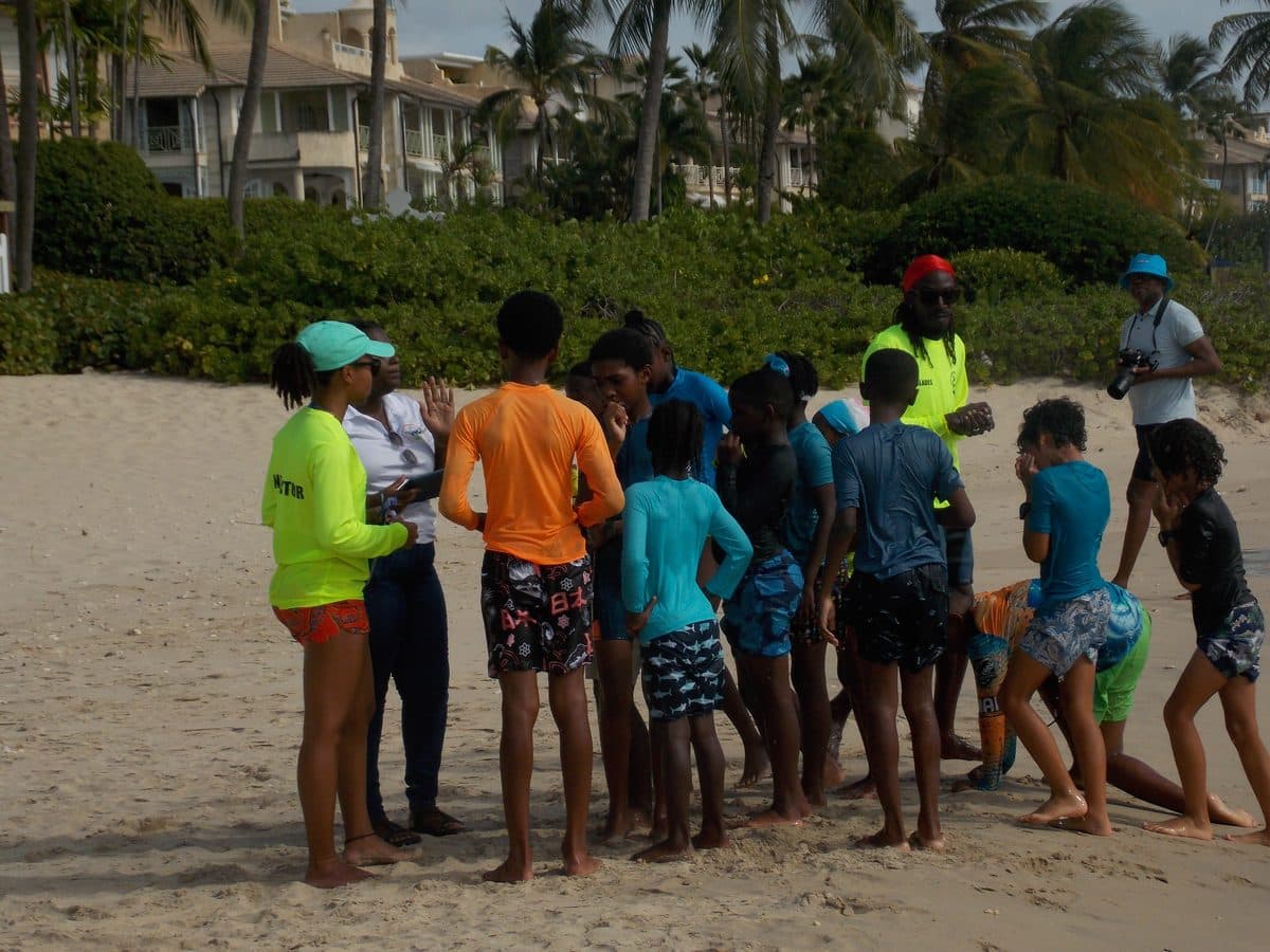 Don Shark Aquatics instructors briefing Mako Club swimmers on the beach, colonial-style clubhouse behind them